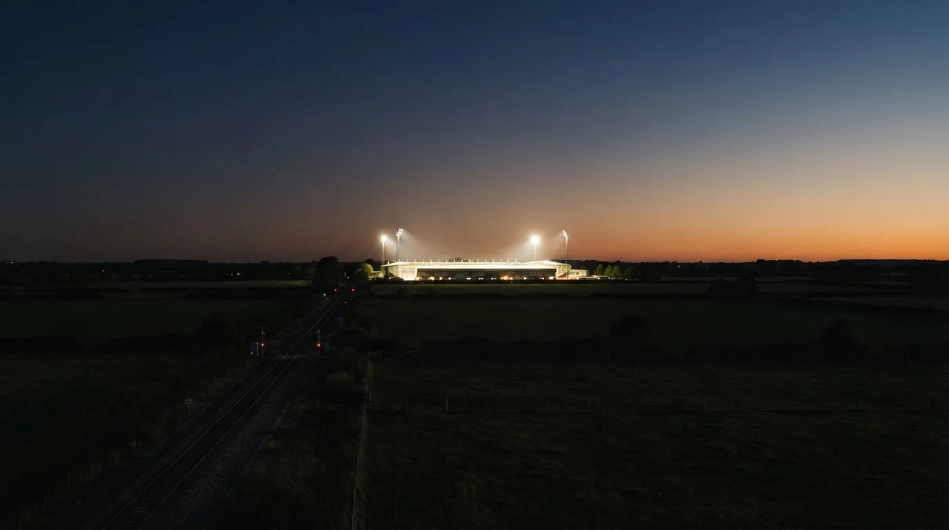 Stadion rozświetlony reflektorami widziany z daleka znad ciemnych pól w pogodny wieczór.