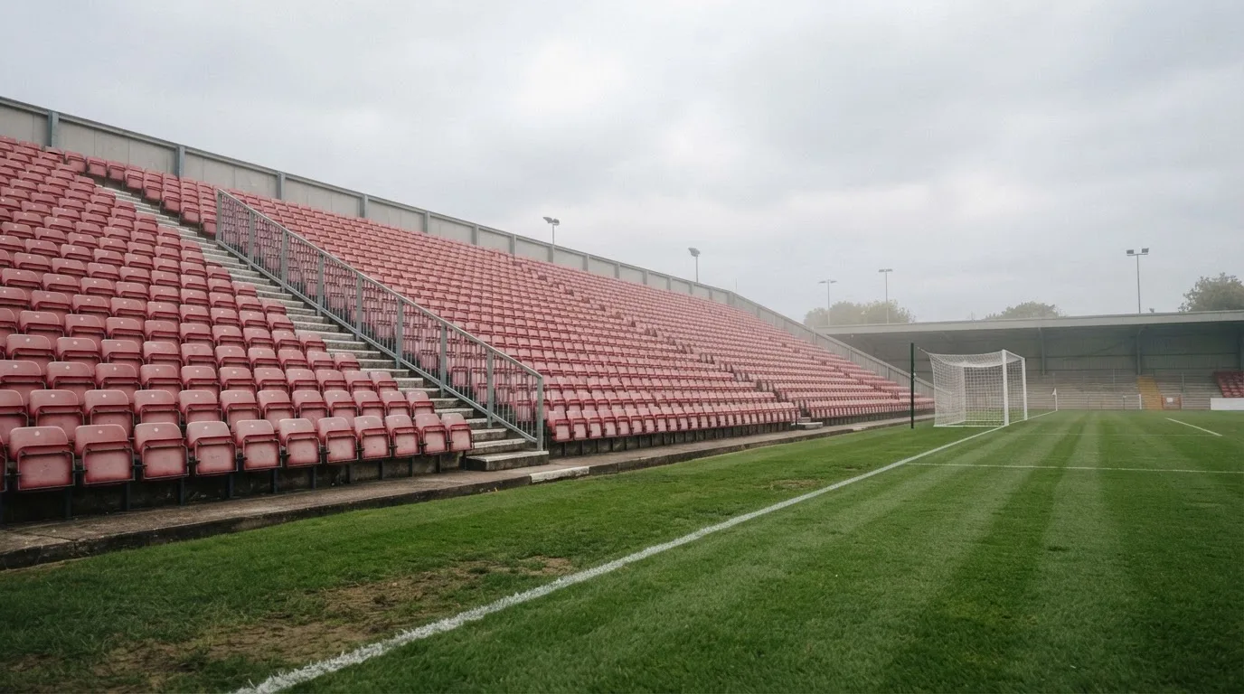 Rzędy pustych plastikowych krzesełek małego stadionu widziane z poziomu murawy pod pochmurnym niebem.